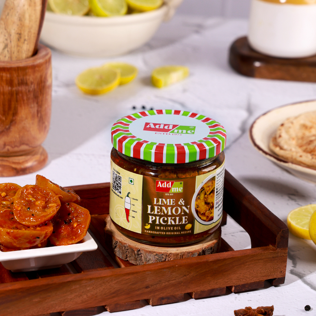 Add Me Foods: Jar of Adda Lime & Lemon Pickle on a wooden tray with lemon slices and snacks in the background.