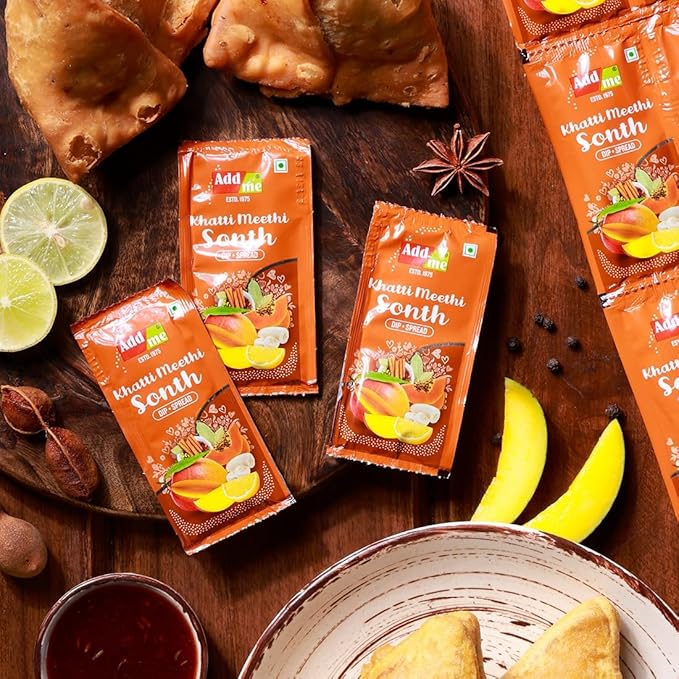 Packages of 'Add Me Khati Meethi Snoth chutney/chatni' on a wooden surface with food items.
