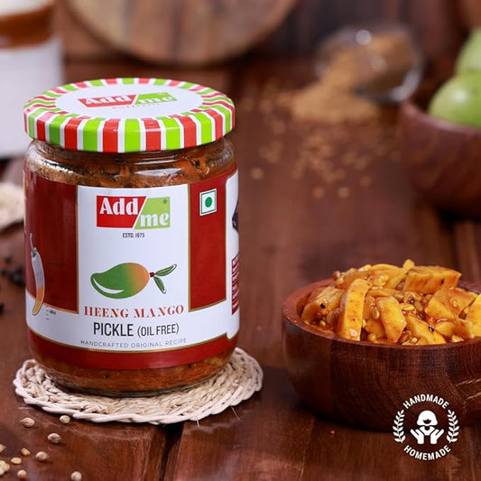 Jar of Add Me Heeng Mango Pickle on a wooden surface with a bowl of pickles.