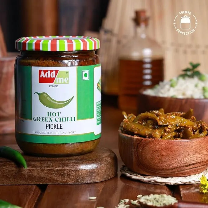 Jar of Add Me hot green chili pickle on a wooden surface with a bowl of pickles in the background.