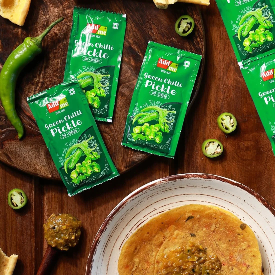 Adda brand green chilli pickle packets on a wooden board with pickles and chilies.