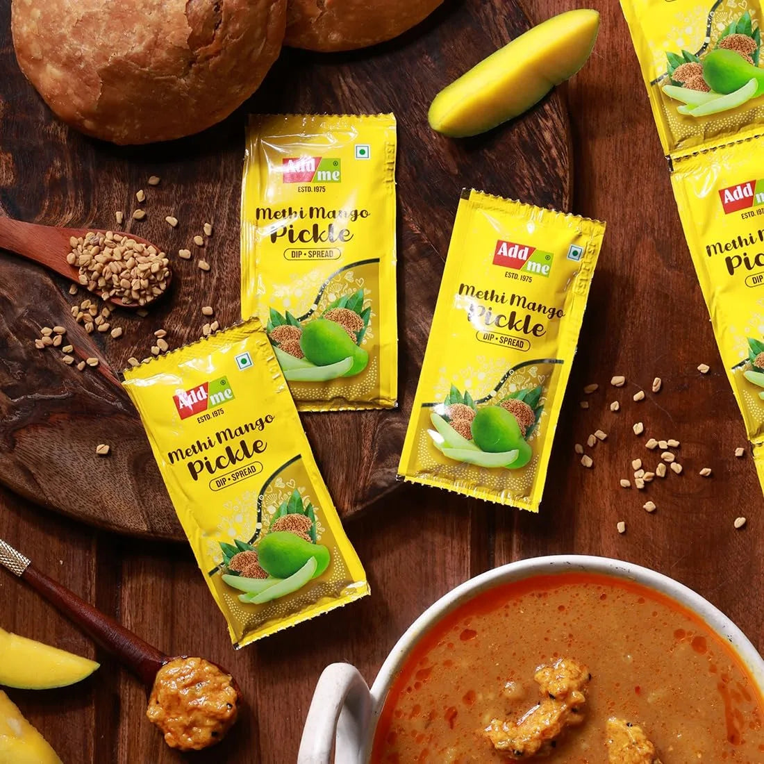 Yellow packets of Add Me Methi Mango Pickle on a wooden surface with ingredients and a bowl of pickle.