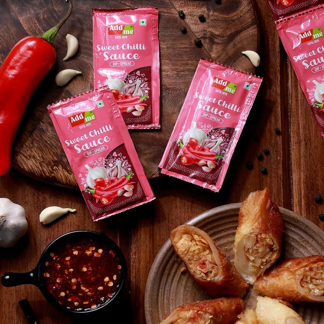 Pink packets of 'Add Me Sweet Chilli Sauce' on a wooden surface with ingredients and food items.