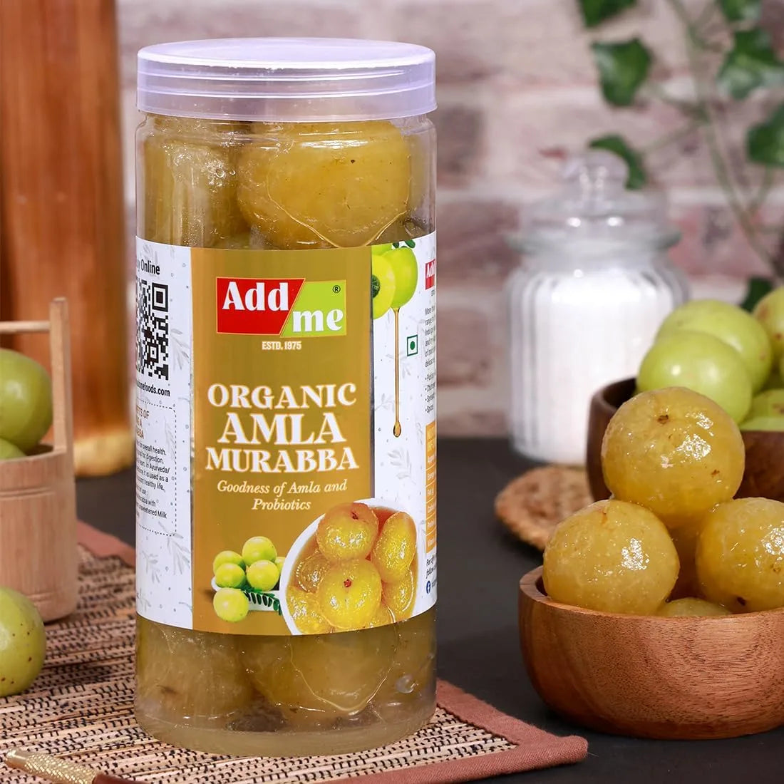 Jar of Add Me Organic Amla Murabba with a label on a table with a wooden bowl of amla berries.