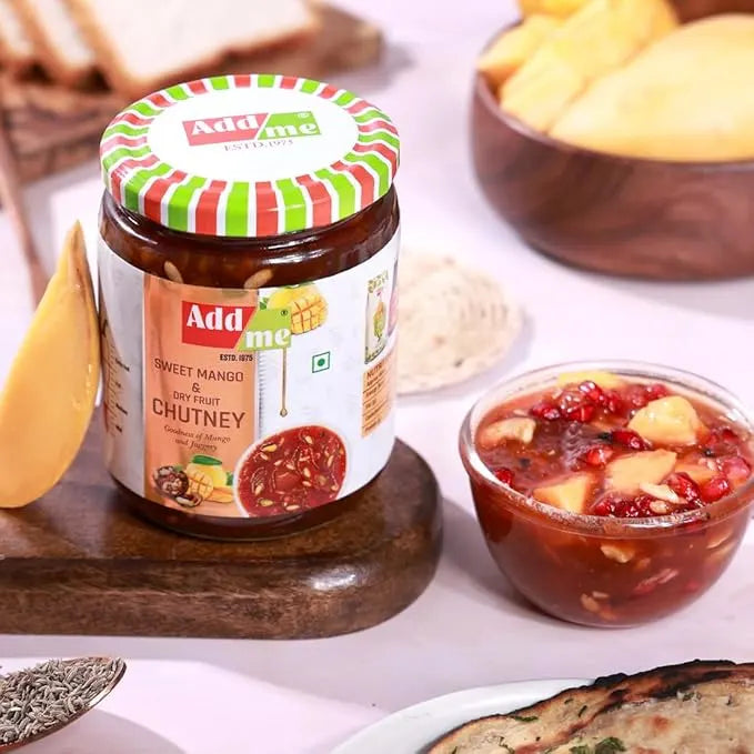 Jar of Add Me Sweet Mango & Dry Fruit Chutney on a wooden board with a bowl of chutney and snacks in the background.