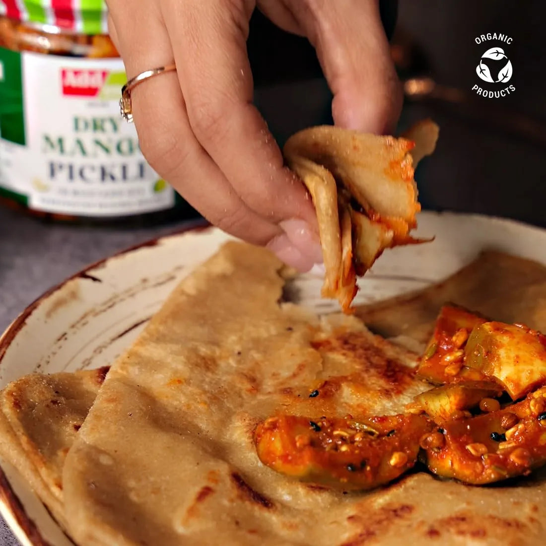 Person dipping a piece of flatbread into a bowl pickle with a jar of dry mango pickle in the background.