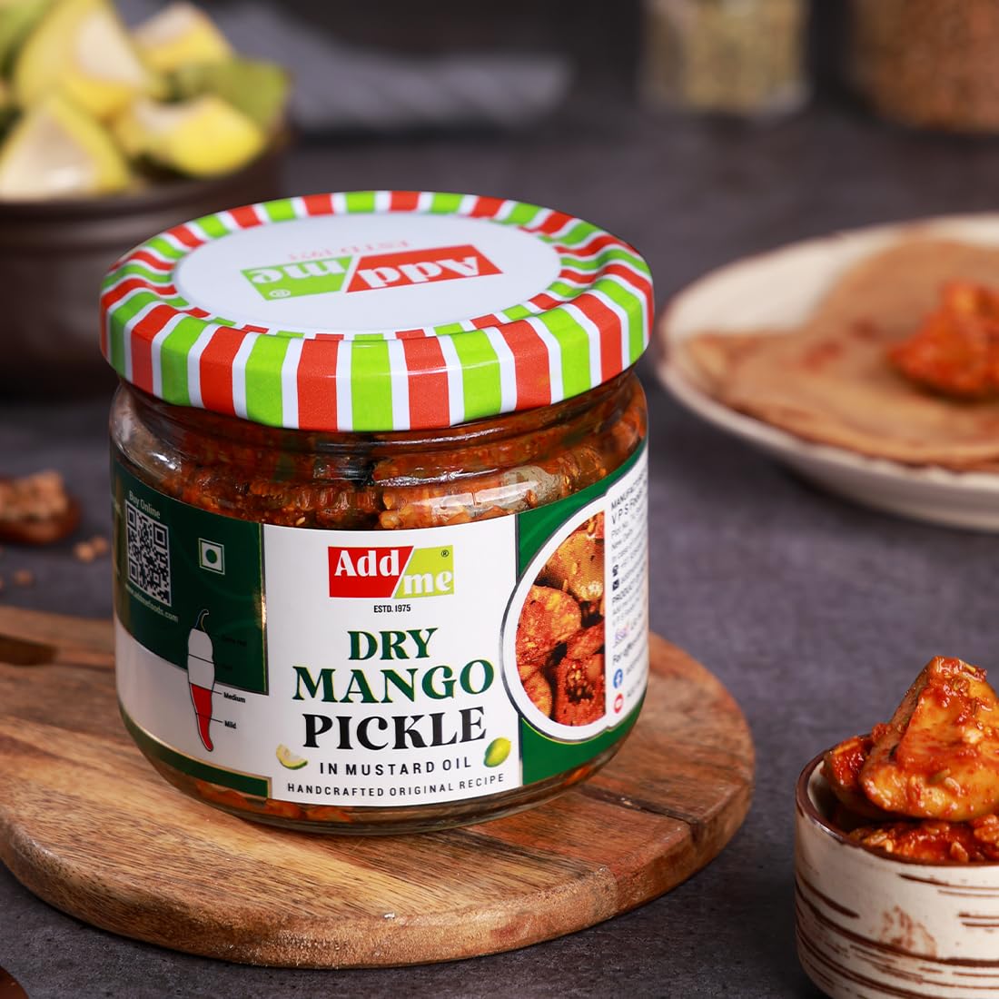 Jar of dry mango pickle with a colorful lid on a wooden board, with food items in the background.