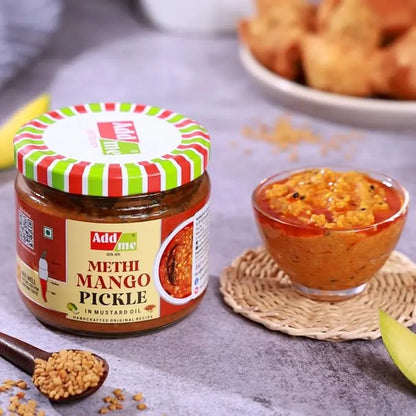 Jar of Add Me Methi Mango Pickle with a small bowl of pickle and spices on a table.
