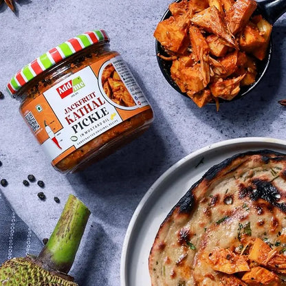 Jar of jackfruit kathal pickle with a plate of flatbread and jackfruit pieces on a gray surface.