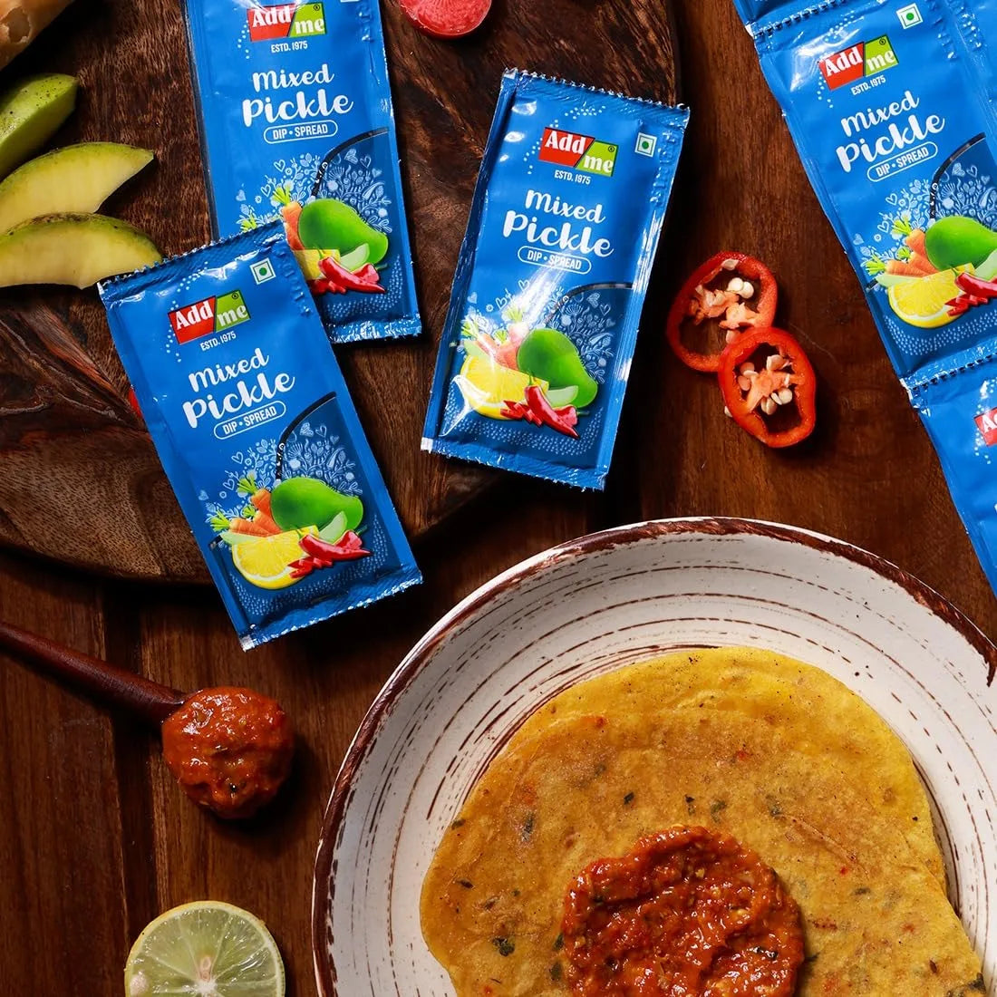 Addmi mixed pickle packets on a wooden surface with a bowl of roti and pickle.