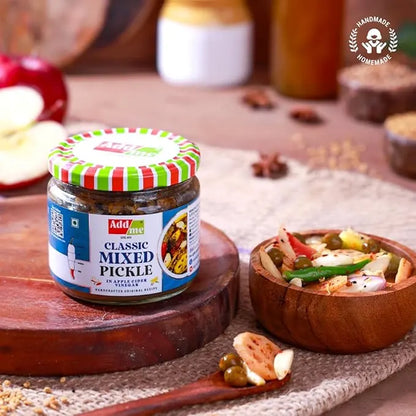 Jar of Adda Classic Mixed Pickle on a wooden board with a bowl of pickles and spices in the background.