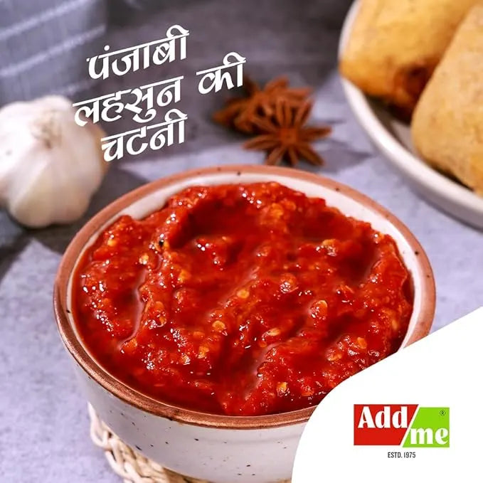 Bowl of red chutney with garlic and bread on a gray surface, featuring Add Me logo.